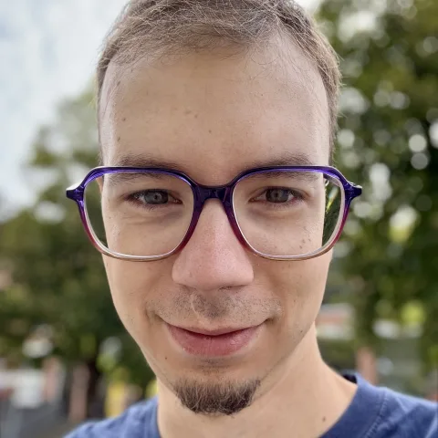 Portrait of Sebastian with purple glasses, smiling in front of green trees